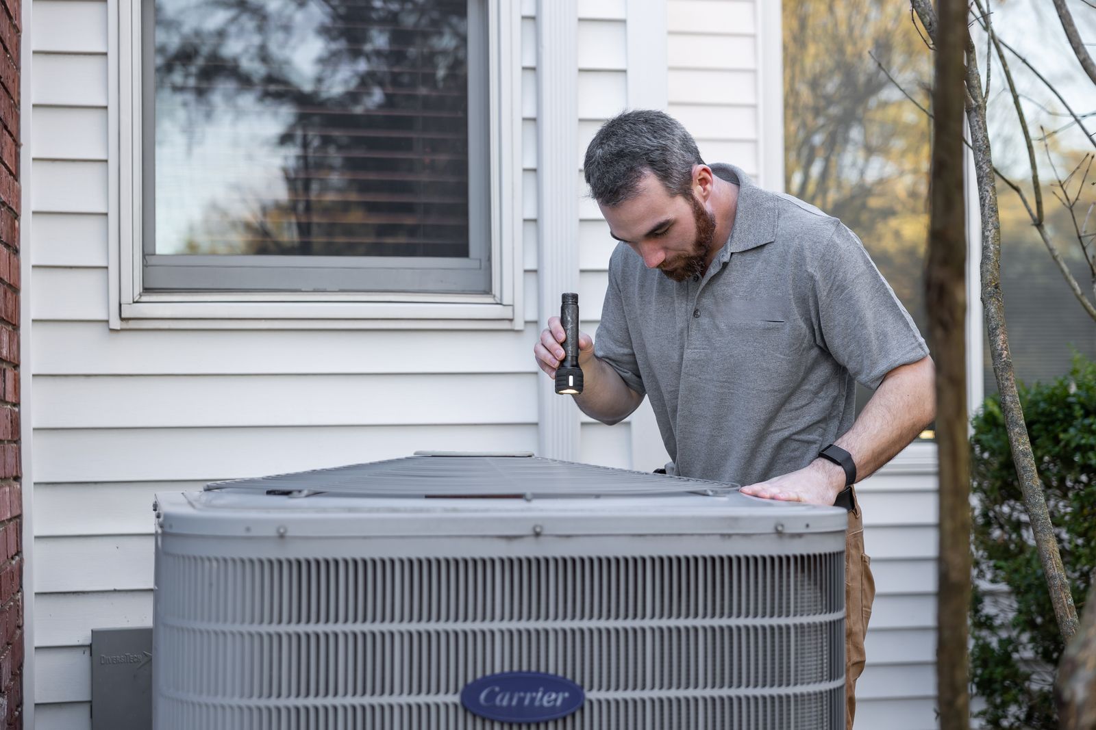 HVAC technician inspecting an outdoor air conditioning unit beside a home with a flashlight